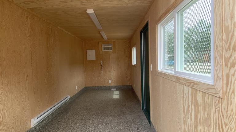 photo shows the interior of a 20' shipping container that has been modified with wood walls, and laminate flooring, baseboard heaters, electrical outlets, 2 caged windows and 1 man door halfway down the right hand side.